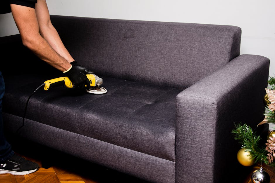 A person wearing a black glove is performing surface cleaning on a gray carpeted floor using a handheld cleaning device attached to a hose, with visible foam and water spray. The carpet appears to be 