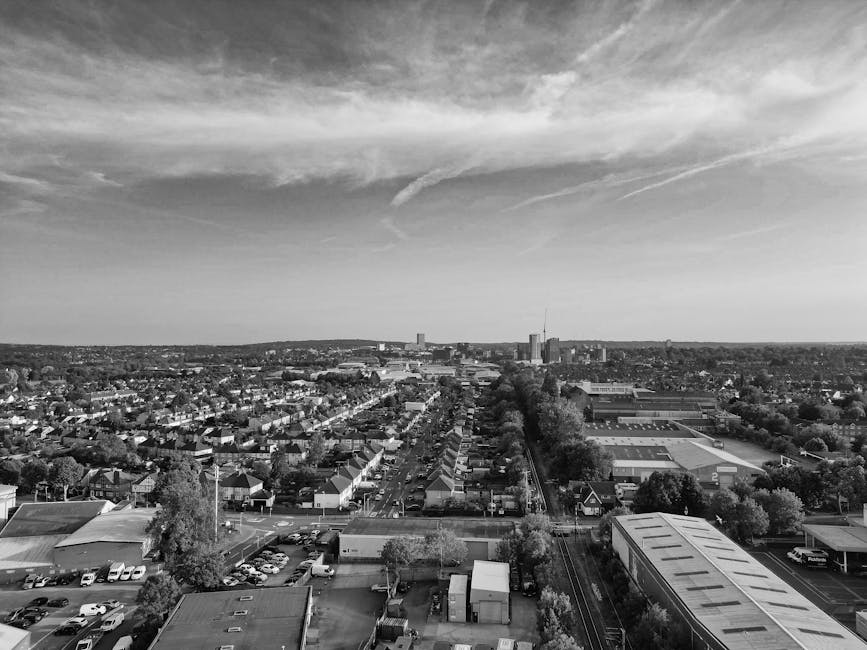 Aerial black-and-white photograph of an industrial and commercial area featuring large warehouse buildings with flat and slightly curved roofs, surrounded by parking lots filled with numerous parked v