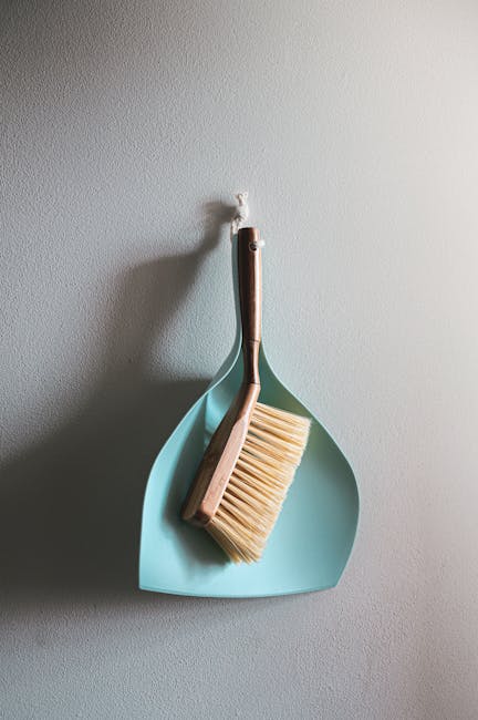 A light blue dustpan and a wooden-handled brush with beige bristles hanging on a white textured wall, arranged with the dustpan facing downward and the brush resting inside it. The scene is evenly lit